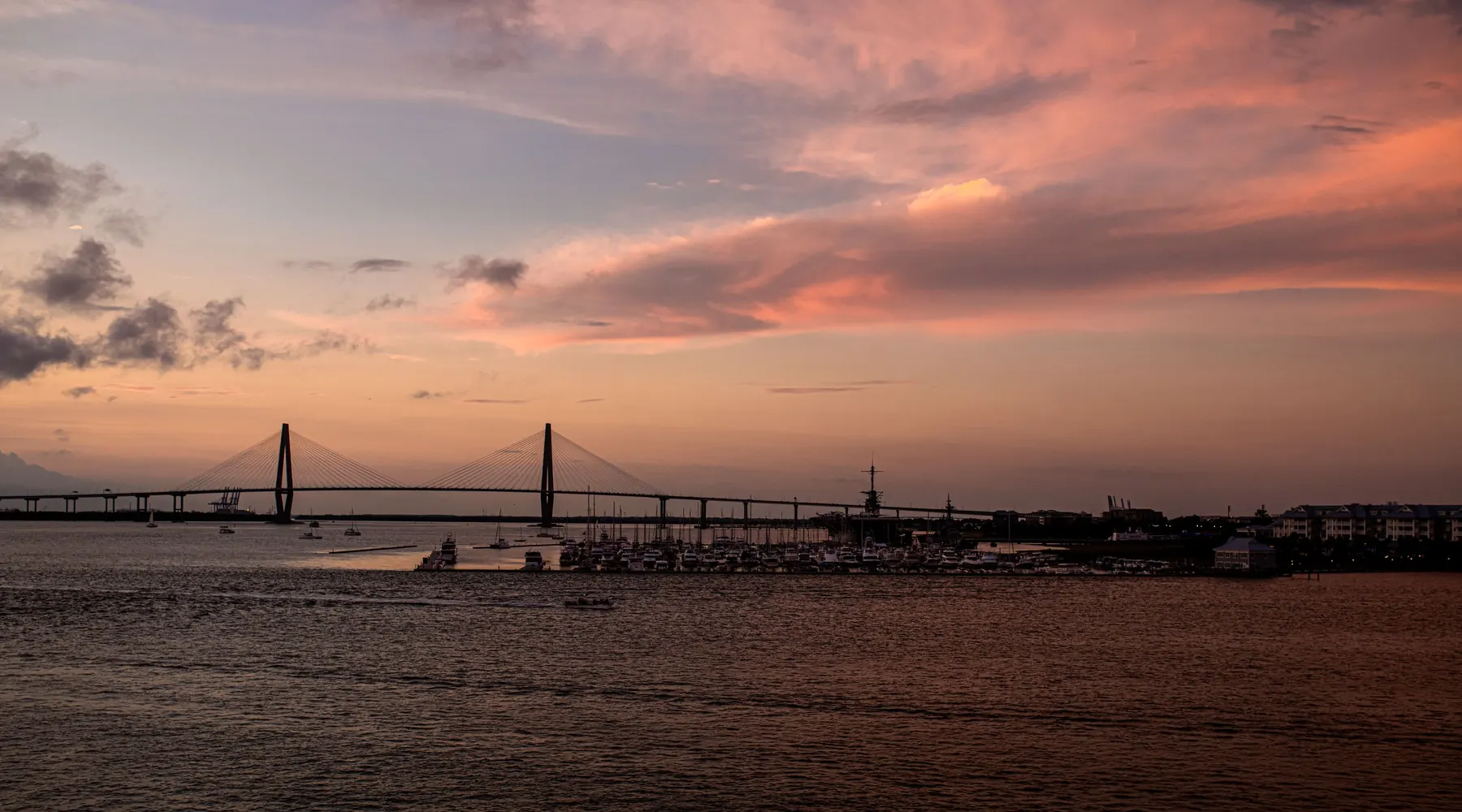 Charleston Harbor at dusk, with the Ravenel Bridge's two towers on the horizon, a marina in silhouette and copper clouds over the water