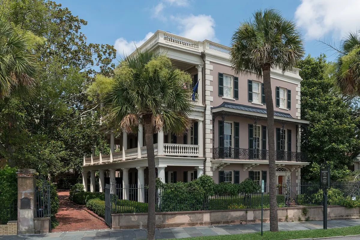A pale pink antebellum house with columned piazzas and a balustrade on East Battery, set behind a wrought-iron fence and framed by palmetto trees