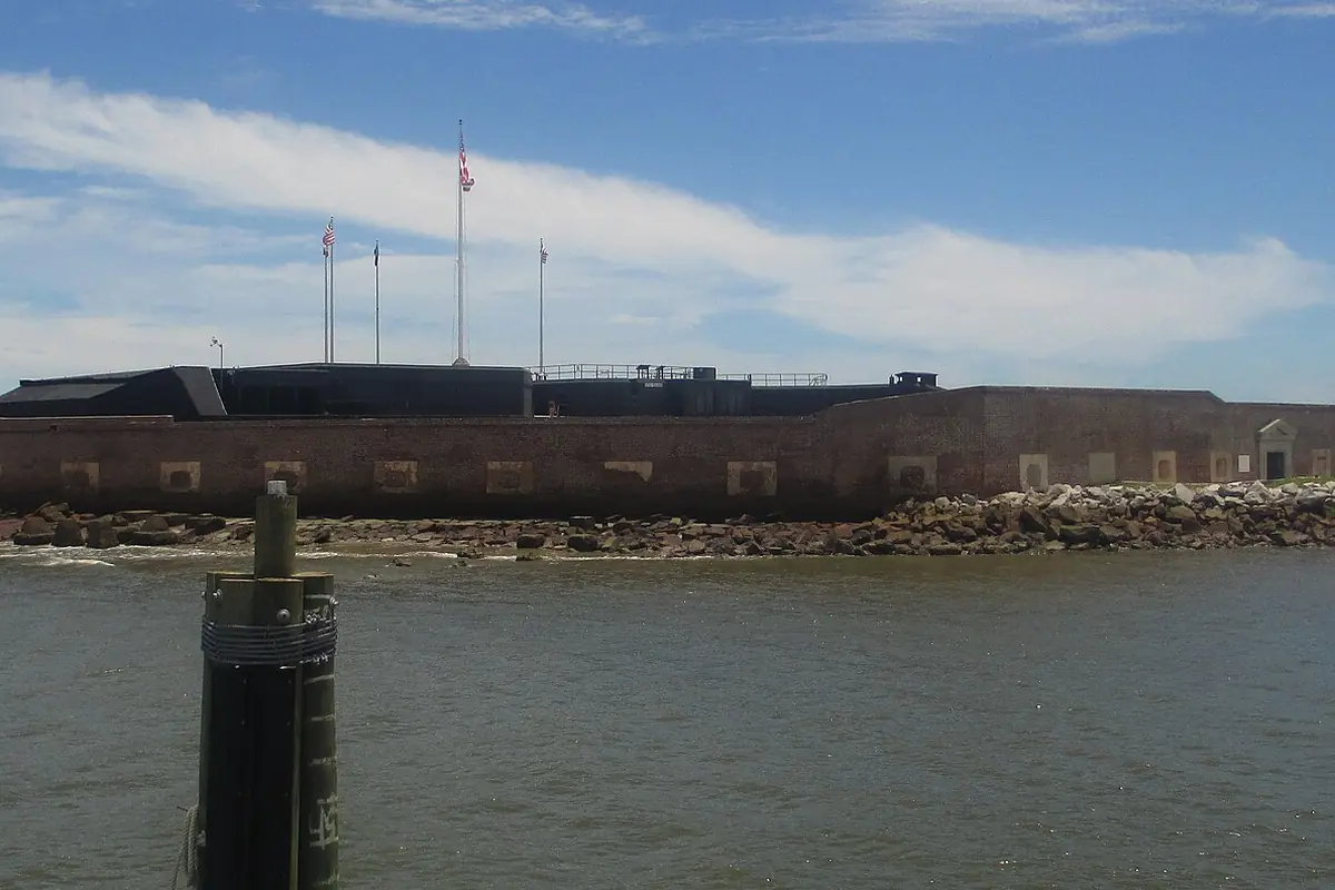The low brick walls of Fort Sumter on its man-made island, with flagpoles above the wall and rocks along the waterline under a blue sky