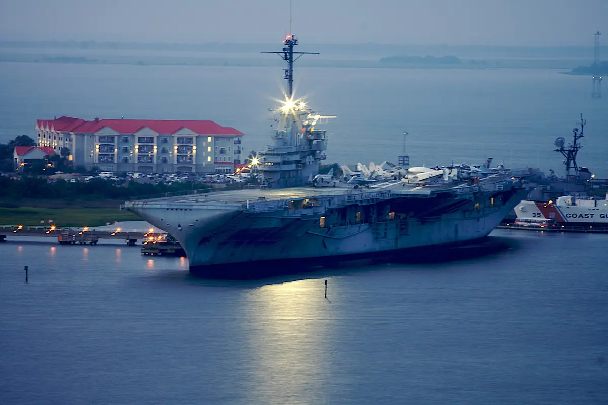 The aircraft carrier USS Yorktown moored at Patriots Point at dusk, its masthead light shining over still water, with a lit hotel with a red roof on the shore beside it