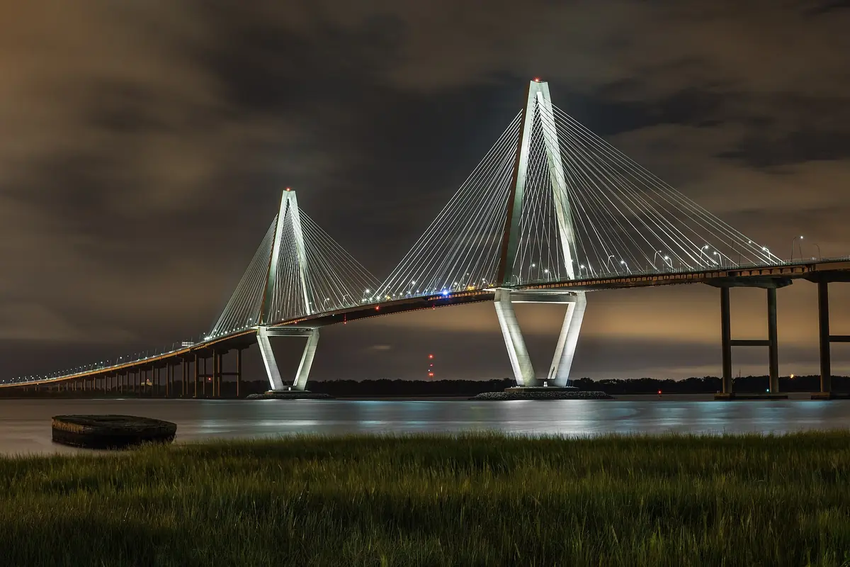 The Arthur Ravenel Jr. Bridge lit at night, its two diamond-shaped towers and fan of cables reflected in the Cooper River, with marsh grass in the foreground