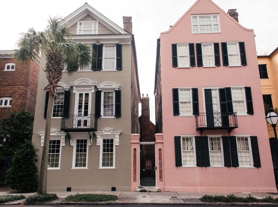 Two tall narrow Charleston houses, one grey and one pink, with black shutters and a palmetto tree between the doorways